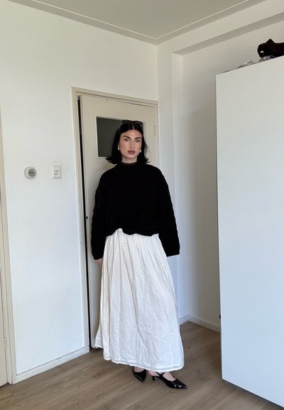 Mujer con cabello negro, vestida con suéter negro, falda larga blanca y tacones negros, de pie en el interior cerca de una puerta cerrada y un armario blanco.
