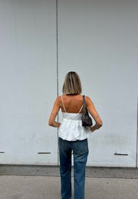 Top de tecido branco com alças finas, design peplum, combinado com jeans de denim azul. A modelo segura uma pequena bolsa escura. O fundo é uma parede cinza lisa.