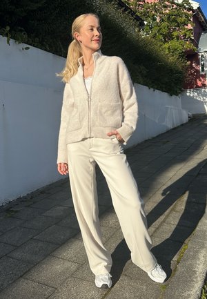 Mujer de pie en una acera iluminada por el sol junto a una pared blanca, vestida con un suéter crema con cremallera, pantalones blancos y zapatillas blancas.