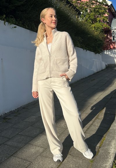 Mujer de pie en una acera iluminada por el sol junto a una pared blanca, vestida con un suéter crema con cremallera, pantalones blancos y zapatillas blancas.