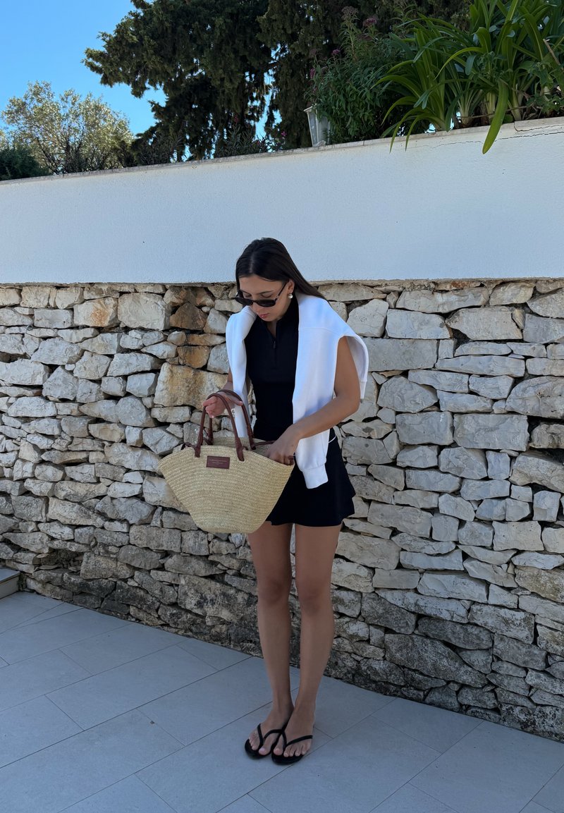 Femme portant une robe noire et des lunettes de soleil tenant un sac à main tressé, debout sur un sol carrelé devant un mur en pierre avec des plantes au-dessus.