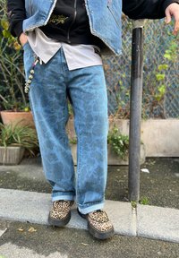 Calças de ganga com um padrão abstrato azul claro, combinadas com sapatos slip-on com padrão de leopardo. Usadas com uma blusa em camadas cinza e preta.