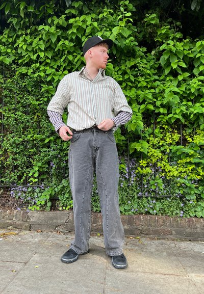 Camisa de manga larga a rayas con jeans gris oscuro y zapatos negros, de pie contra un follaje verde, mostrando un atuendo urbano casual.