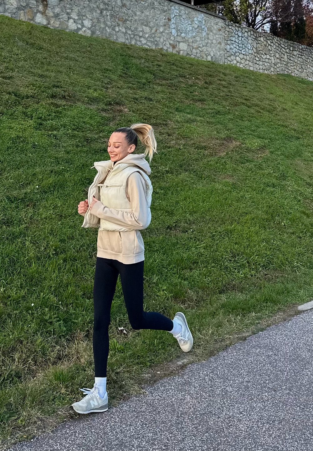 Un gilet beige sur un sweat à capuche clair, des leggings noirs et des chaussures de course claires. Herbe et un mur en pierre en arrière-plan.