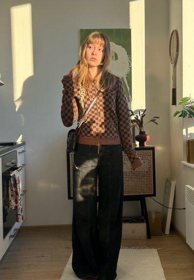 Joven mujer con sudadera de cuadros y pantalones anchos negros de pie en una cocina iluminada por el sol con un armario vintage, plantas y arte en la pared.