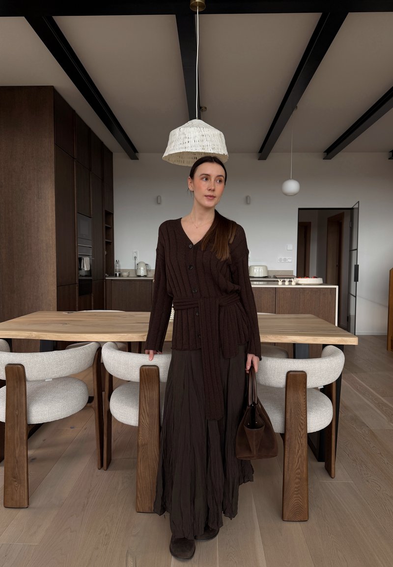 Femme en cardigan marron foncé et jupe longue se tient dans une cuisine moderne en bois, tenant un sac à main marron près d'une table à manger avec des chaises blanches.