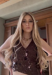 Jeune femme aux longs cheveux blonds portant des lunettes, un haut marron sans manches à sequins et des colliers superposés, debout à l'intérieur les mains sur les hanches.