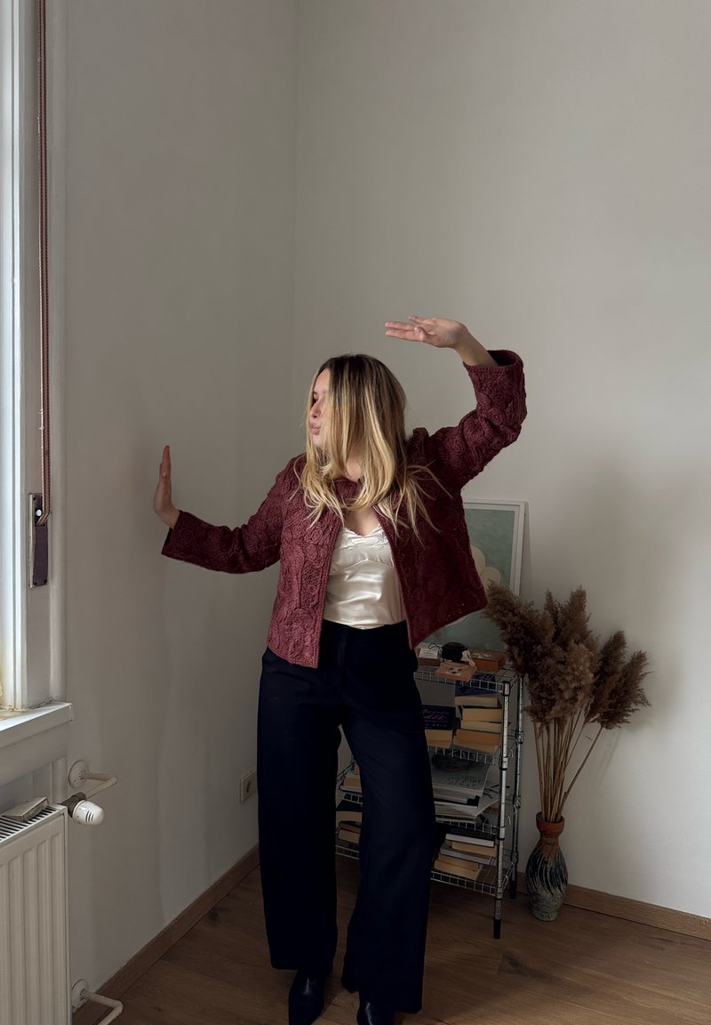 Femme adoptant une pose dynamique près d'une fenêtre, portant une veste bordeaux texturée, un chemisier crème et un pantalon noir dans une pièce minimaliste avec des livres et des plantes séchées.