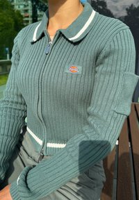 Grønn ribbestrikket cardigan med glidelås foran, hvit stripete krage og logo-patch på brystet. Har tettsittende ermer og teksturert stoff.