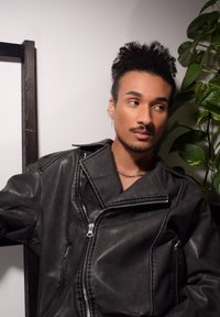 Veste en simili cuir noir avec un large col et une fermeture à glissière asymétrique, dotée d'une poche et de détails de couture. Chaîne en argent visible.