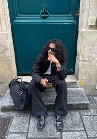 Mężczyzna z kręconymi włosami i okularami przeciwsłonecznymi siedzi na kamiennym stopniu, trzymając telefon, ubrany na czarno, obok niego leży czarna torba.