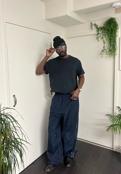 Pantalones ligeros azul marino con rayas blancas a los lados, combinados con una camiseta negra ajustada y un gorro negro, complementados con gafas transparentes.