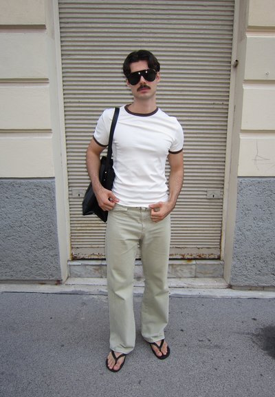 Camiseta blanca con ribetes marrones, pantalones verde claro, chanclas negras y un bolso negro de hombro. Lleva gafas de sol y un bigote.