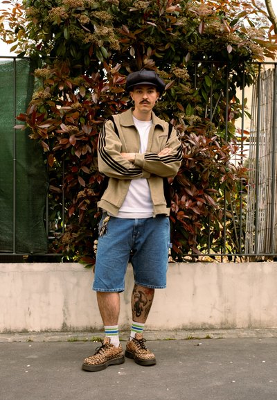 Hombre con bigote permanece de pie con los brazos cruzados frente a arbustos, lleva una gorra, chaqueta beige, camisa blanca, pantalones cortos vaqueros y zapatos con estampado de leopardo.