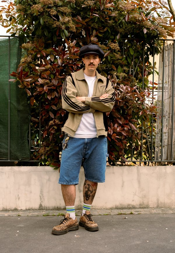 Hombre con bigote permanece de pie con los brazos cruzados frente a arbustos, lleva una gorra, chaqueta beige, camisa blanca, pantalones cortos vaqueros y zapatos con estampado de leopardo.