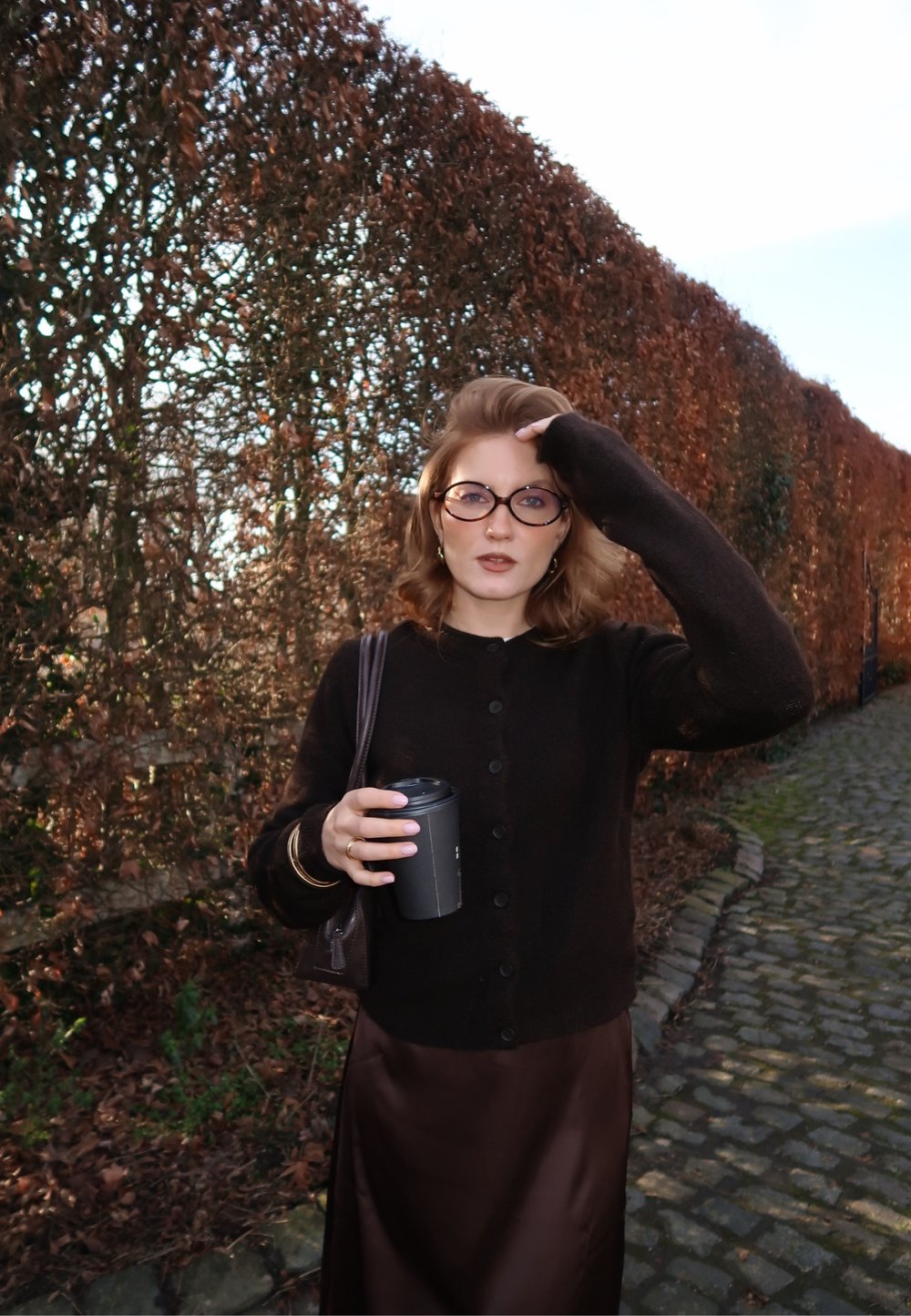 Femme portant des lunettes et des vêtements foncés tenant une tasse de café, debout sur un chemin pavé à côté d'une grande haie aux feuilles brunes.