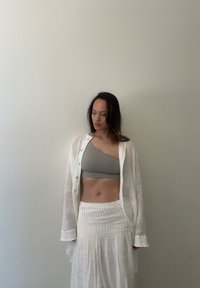 Camisa blanca de lino de manga larga sobre un sujetador gris de un hombro y una falda blanca texturizada. La tela parece ligera y fluida.