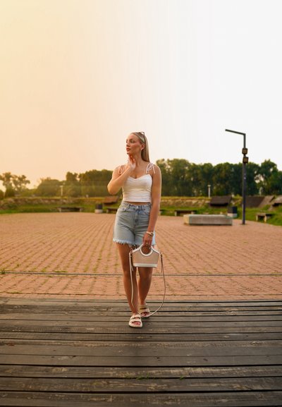 Top blanco sin mangas con tirantes de encaje, pantalones cortos de mezclilla y bolso beige con asa redonda. De pie sobre tablas de madera en un entorno al aire libre.