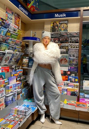 Una modelo vestida con una chaqueta de piel blanca esponjosa, pantalones de chándal grises y tacones blancos, está de pie junto a un estante de revistas etiquetado como "Computadora" y "Música".