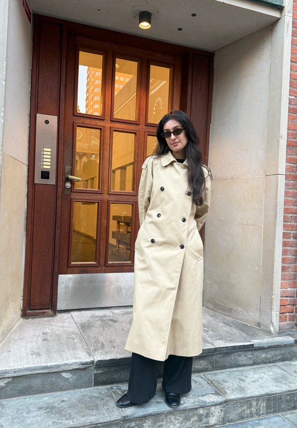 Una mujer con gafas de sol y una gabardina beige está de pie en unos escalones, frente a una entrada de madera con paneles de vidrio.