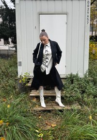 Svart lång kappa över en vit skjorta med svart korssnörning, tillsammans med knähöga vita stövlar. Grön gräs och trätrappor i bakgrunden.
