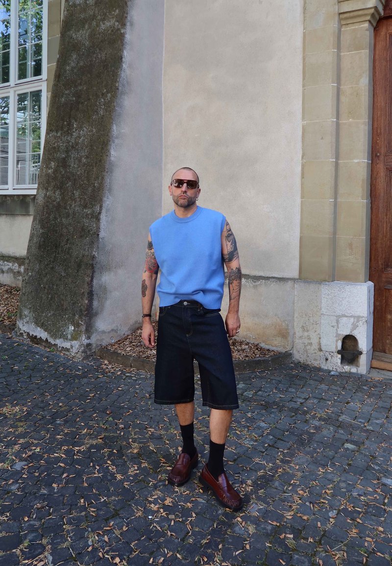 Homme avec des tatouages portant un haut sans manches bleu, des shorts longs foncés, des chaussettes noires et des chaussures marron, debout dans une rue pavée près d'un bâtiment.