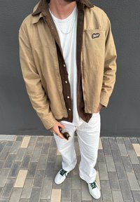 Beżowa kurtka płócienna z brązowym kołnierzem z sztruksu, biała koszulka, białe spodnie, trzyma okulary przeciwsłoneczne; codzienny strój z zielonym akcentem na butach.