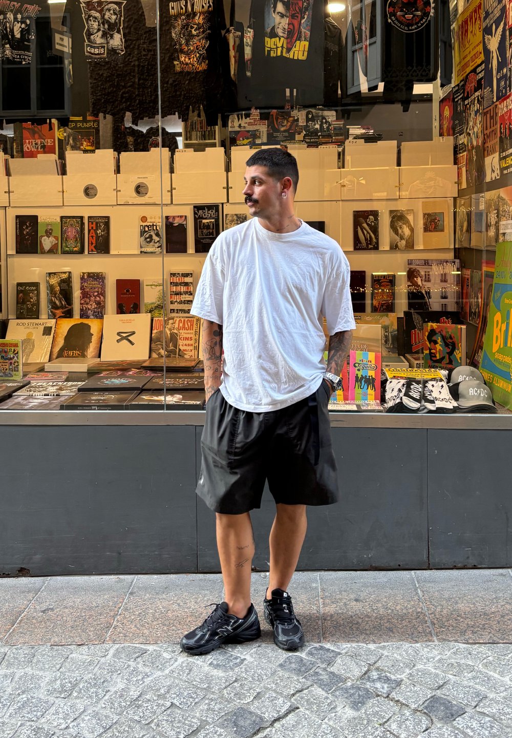 Shorts negri și un tricou alb larg, purtate cu adidași negri, stând în fața unei vitrine de magazin care afișează diverse obiecte legate de muzică.