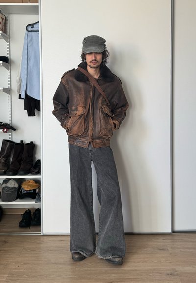 Persona de pie en el interior, vistiendo una chaqueta de cuero marrón desgastada, pantalones anchos grises, una gorra gris y un bolso cruzado, con estantes de botas y sombreros cerca.