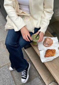 Ένα άτομο σε κρεμ κασκόλ κάθεται σε μια ξύλινη παγκάκι, κρατώντας ένα διάφανο φλιτζάνι πράσινου ποτού, με κρουασάν σε λευκές συσκευασίες κοντά.