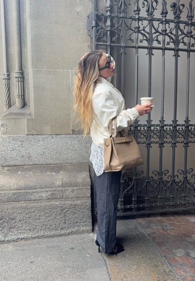Mujer con pelo largo ombré, lleva gafas de sol, chaqueta blanca, pantalones oscuros, sostiene una taza de café y un bolso beige, está de pie junto a una puerta metálica ornamentada.
