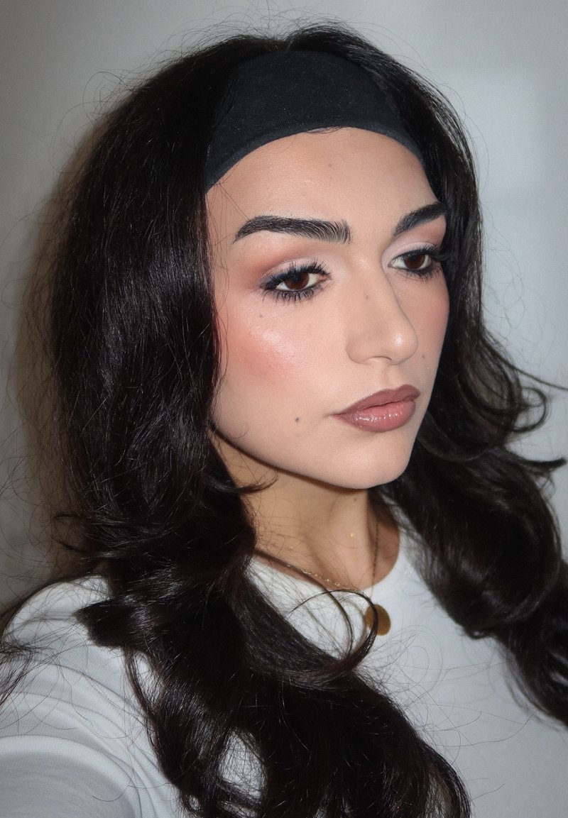 Portrait of a person with long, wavy black hair, wearing a black headband and a white top. Features include natural makeup and a neutral lip color.
