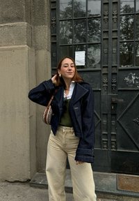 Dark blue denim jacket with a wide collar, a white shirt underneath, a green sweater, and beige wide-leg trousers. Brown patterned handbag.