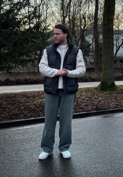 Chaleco acolchado negro sobre un suéter de punto blanco, combinado con pantalones grises holgados y zapatillas blancas. Ambiente natural con árboles de fondo.