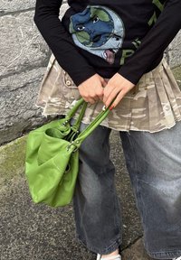 Osoba u crnoj majici s dugim rukavima s grafičkim motivom, maskiranoj plisiranoj suknji preko širokih sivih traperica, držeći zelenu torbu s obojanim noktima.