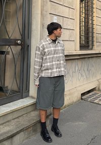 Beige és barna kockás ing, laza szürke rövidnadrággal és fekete fűzős cipővel párosítva. Kültéri helyszín, közel egy kőfalhoz.