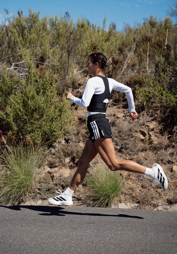 Corredor con pantalones cortos negros, camiseta blanca de manga larga y zapatillas blancas a medio paso en una carretera pavimentada con arbustos secos y cielo azul de fondo.