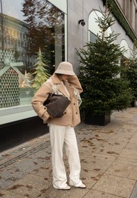 Béžový dvojradový kabát s čipkovaným detailom, krémový top, biele široké nohavice a sivá kabelka; na hlave svetlosivý klobúk v tvare vedra. Pozadie s vianočnou výzdobou.