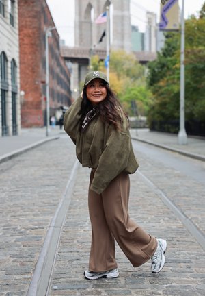 Chaqueta corta verde oliva, pantalones marrones de pierna ancha y zapatillas grises. Lleva una gorra de béisbol y un pañuelo con estampado, situado en una calle empedrada.