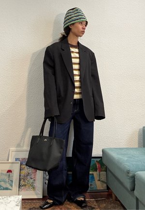 Persona con blazer negro de gran tamaño, camisa a rayas, vaqueros anchos oscuros, gorro multicolor de punto, sosteniendo una bolsa de mano negra, de pie en interiores junto a una obra de arte.