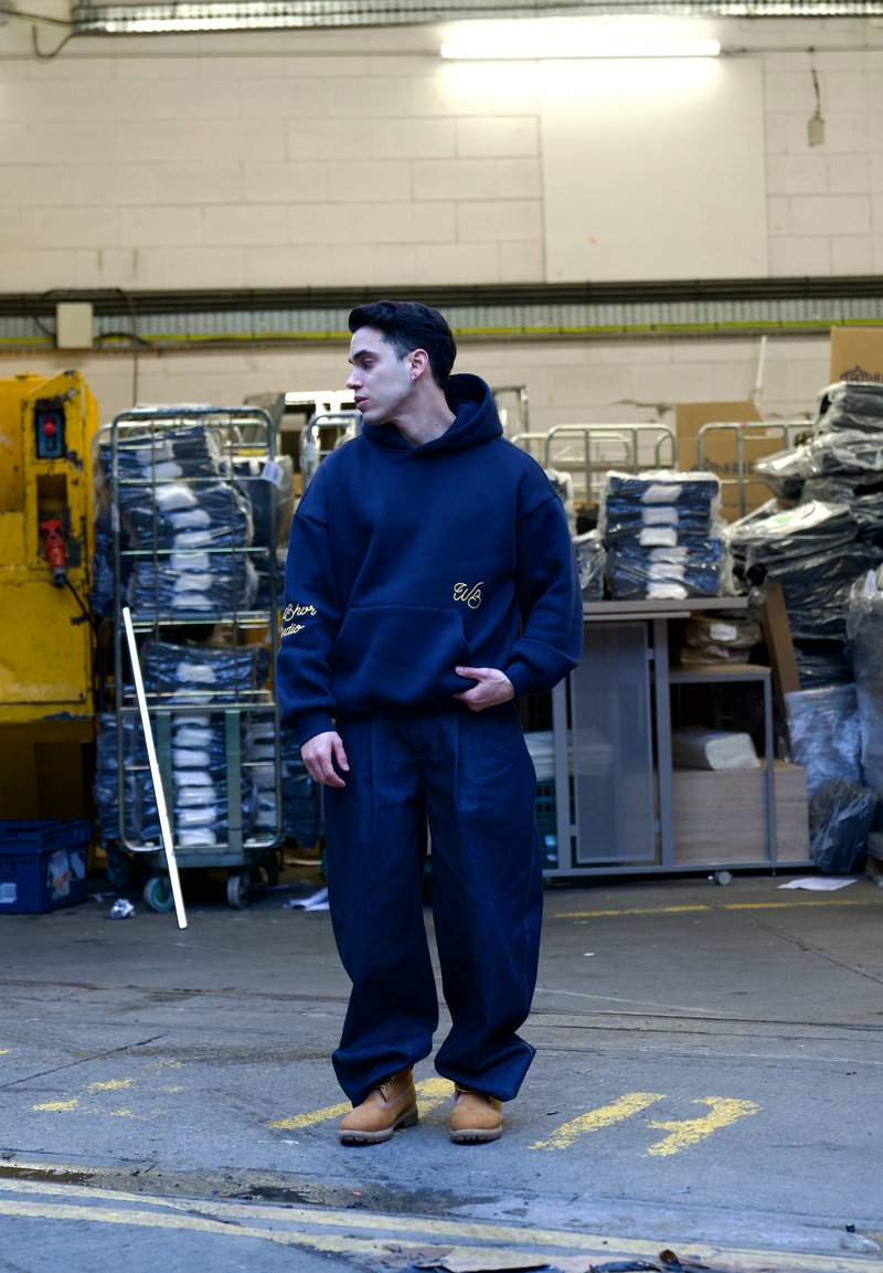 Marineblaue übergroße Kapuzenjacke mit gestickten Details, kombiniert mit dunklen Jeanshosen mit weitem Bein und braunen Clogs, vor einem industriellen Hintergrund.