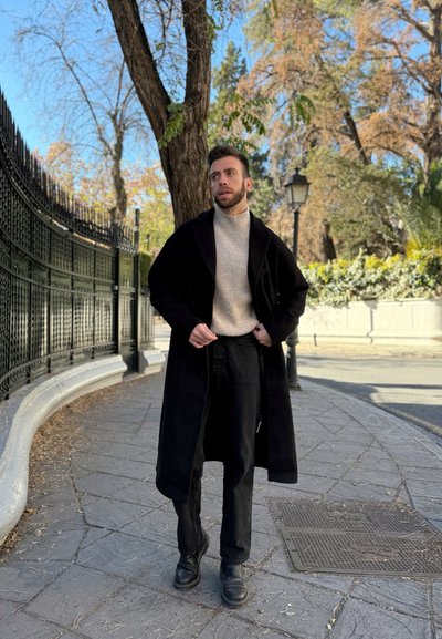 Abrigo largo negro sobre un suéter beige, combinado con pantalones negros y botas de tobillo. De pie en una pasarela de piedra texturizada cerca de árboles.