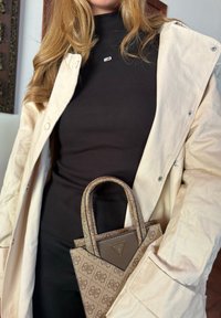 Borsa a tracolla con motivo marrone chiaro e un accento triangolare marrone. Il modello indossa un cappotto beige sopra un dolcevita nero a coste.