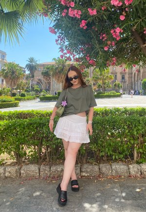 Modelo vistiendo una camisa oversized de color verde oliva y una falda blanca con pliegues, combinada con sandalias de plataforma negras y un pequeño bolso verde con un acento de flor.