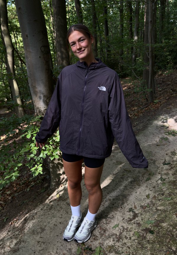 Giovane donna che indossa una giacca scura, pantaloncini neri, calzini bianchi e scarpe da ginnastica, sorridente su un sentiero nel bosco con alberi sullo sfondo.
