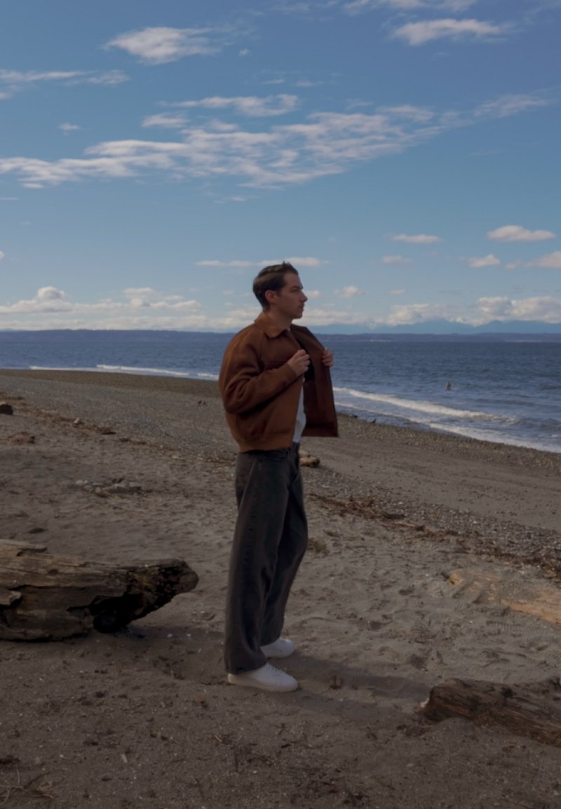 Ein Mann in brauner Jacke und dunkler Hose steht an einem Sandstrand, hält die Jackenrevers und blickt nach rechts zum Ozean unter einem teilweise bewölkten Himmel.