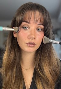 Scène d'application de maquillage mettant en vedette une personne aux longs cheveux ondulés, tenant des pinceaux de maquillage près de son visage, portant des tons de maquillage doux et naturels.