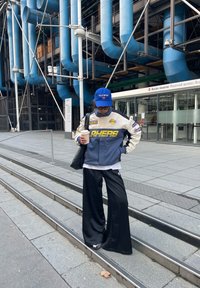 Veste bleue et crème avec texte jaune, pantalon large noir, et casquette bleue. Tient une tasse de café et se tient sur des escaliers extérieurs.