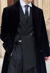 Manteau en velours noir associé à un costume noir, une chemise blanche et une cravate noire. Comprend un design croisé et un pantalon plissé.