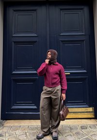 Person wearing a burgundy jumper, brown hood, loose brown trousers, and holding a brown handbag stands in front of large dark blue doors.